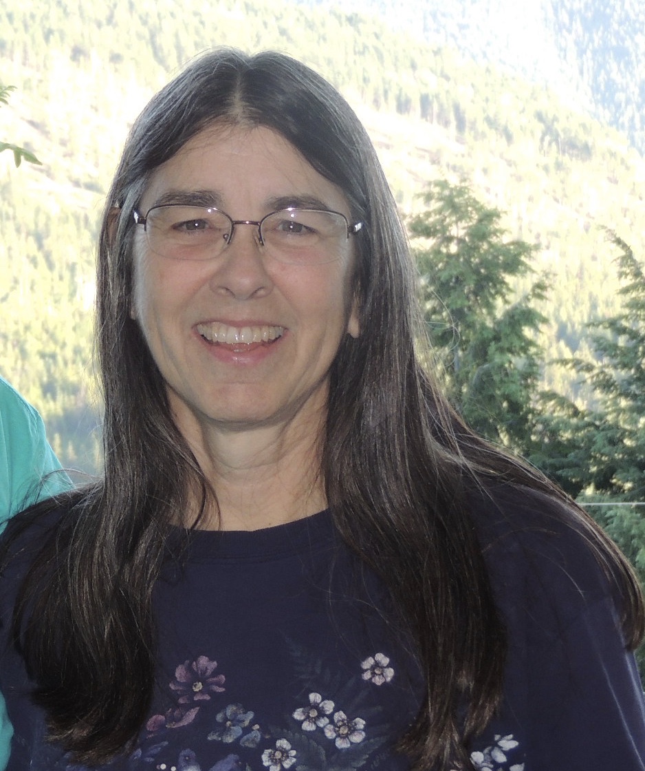 Mindy Smith smiles into the camera. They have long black and white hair and are wearing a purple floral shirt and glasses.